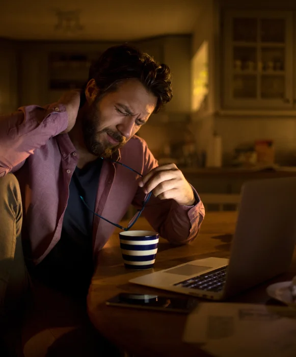 A man at a laptop late in the evening, wincing and holding his stiff neck